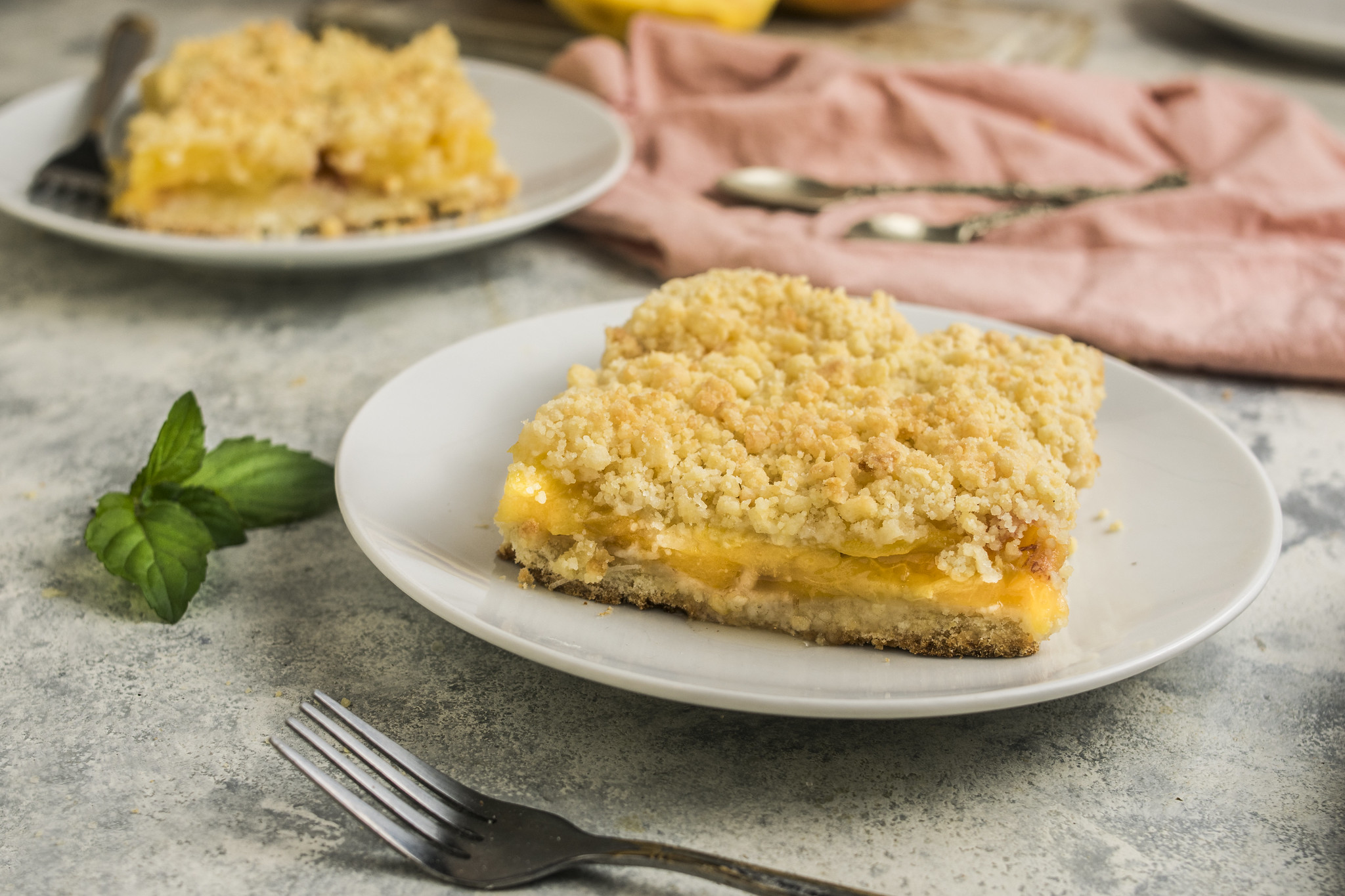 Slice of sbriciolata di pesche served on a white plate, with crumbly topping, soft peach filling and a second slice blurred in the background.